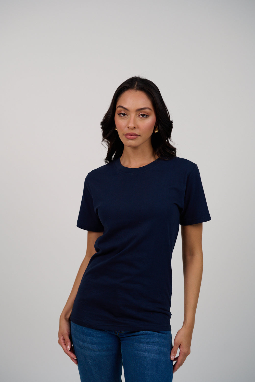 Woman with long dark hair wearing a plain white t-shirt and dark jeans standing against a light grey background. She is facing forward with a slight smile and one hand in her pocket.