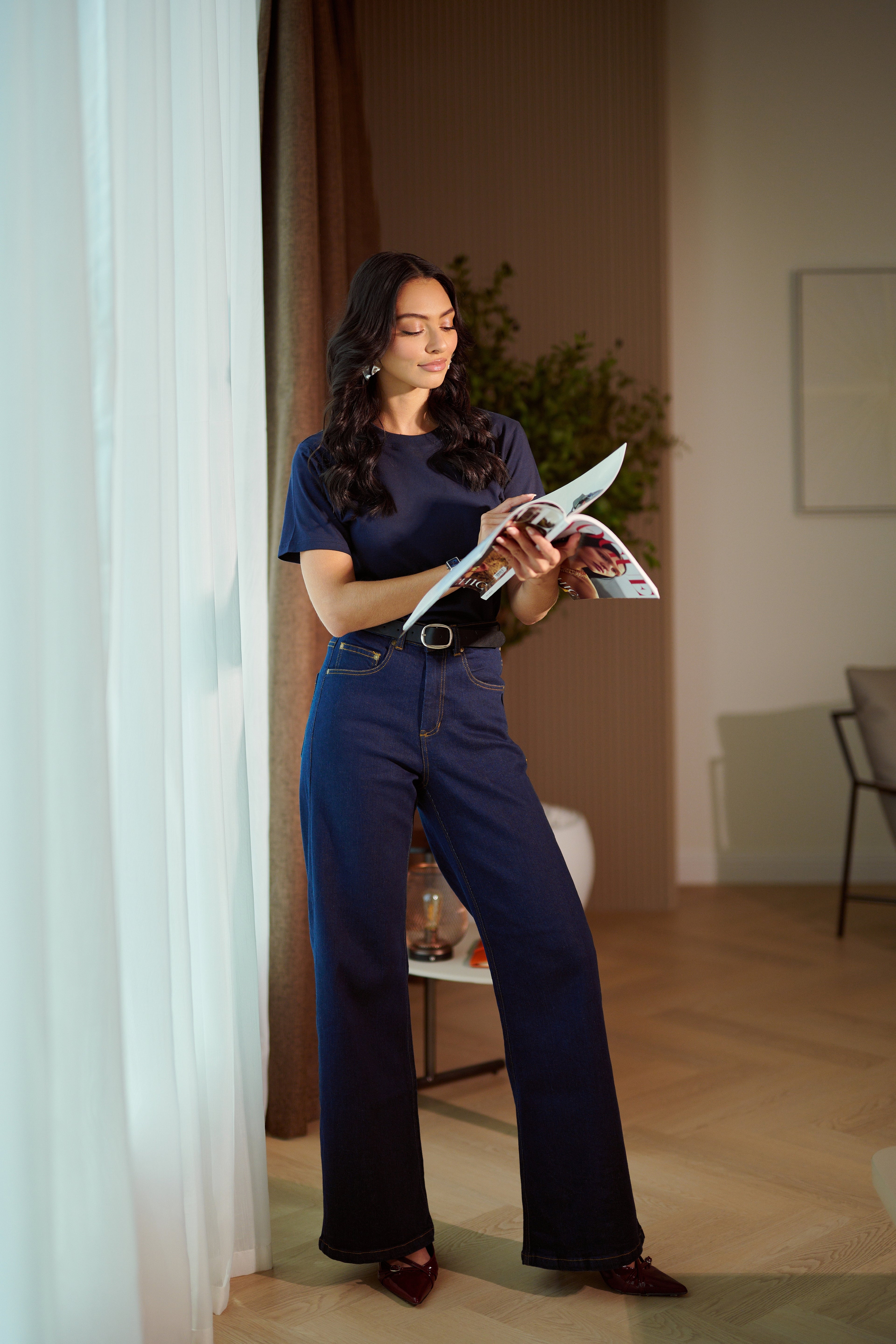 Woman holding a magazine in a room with a plant and chair in the background