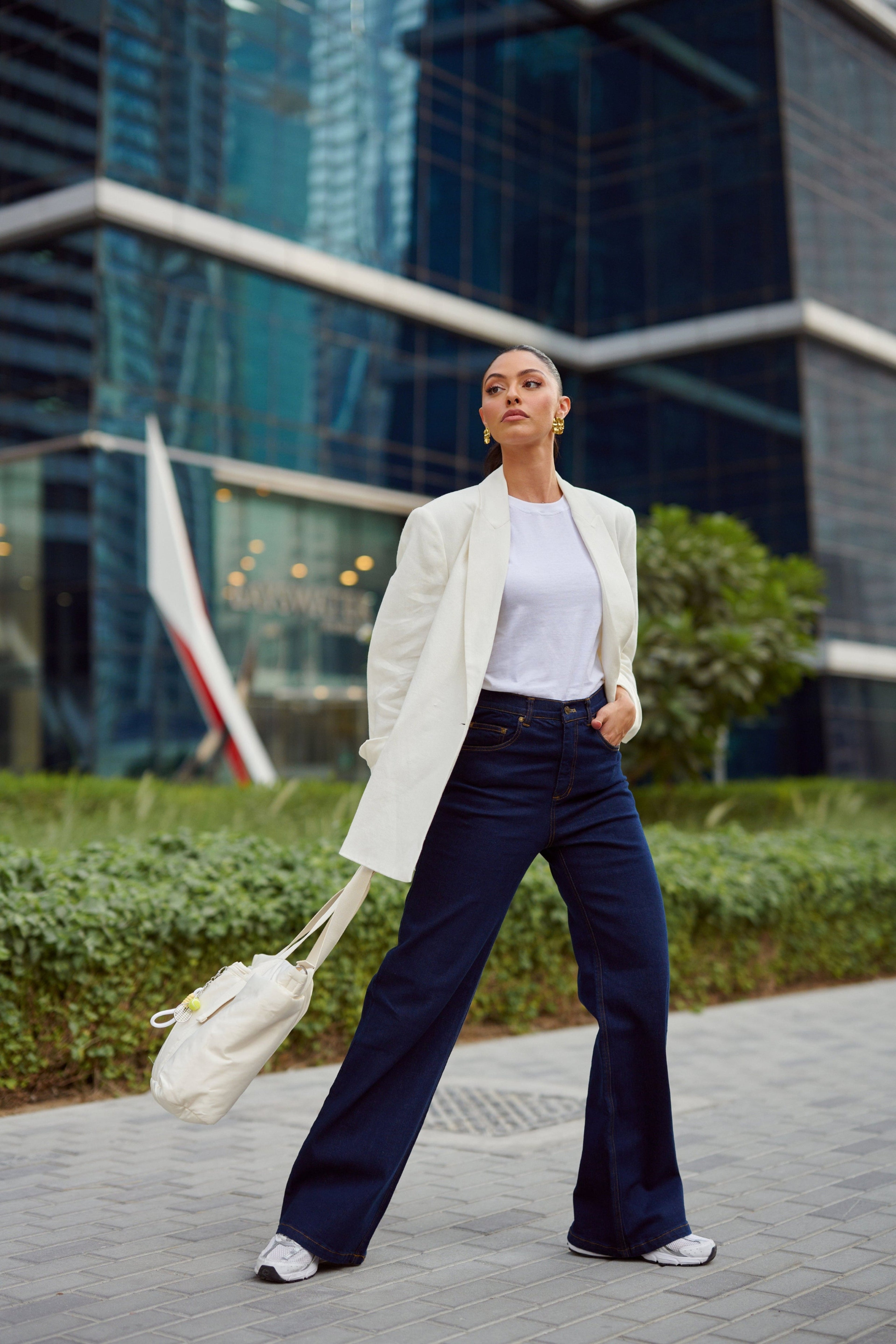 Woman in a white coat and dark pants walking in an urban setting with modern buildings in the background.