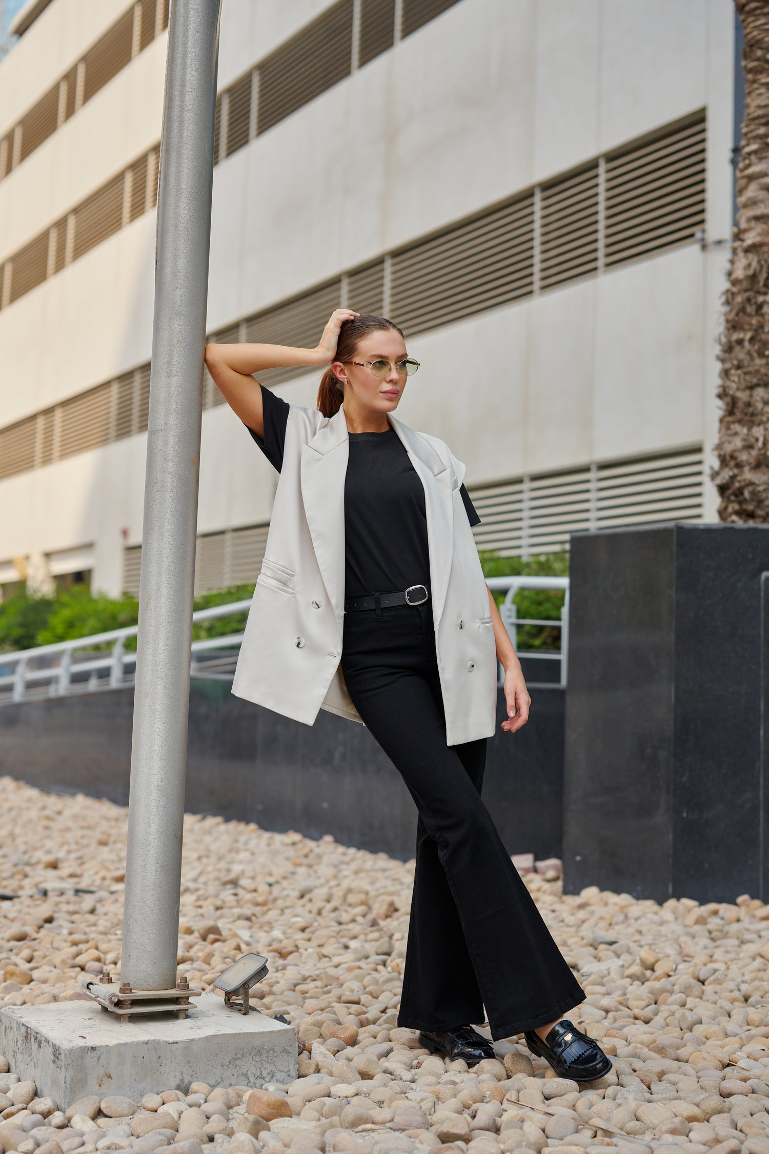 Woman in a black outfit with a white jacket standing on pebbles near a building.
