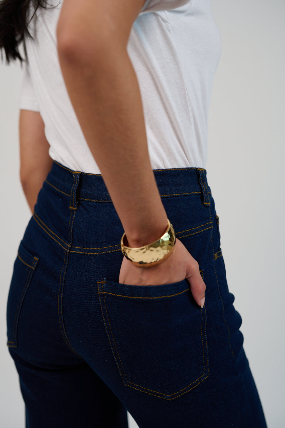 Person wearing blue jeans and a white shirt with a gold bracelet on a plain background