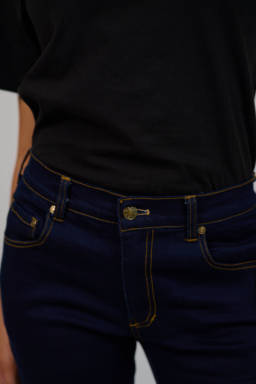 Person wearing a black t-shirt and dark blue jeans with gold stitching on a plain background